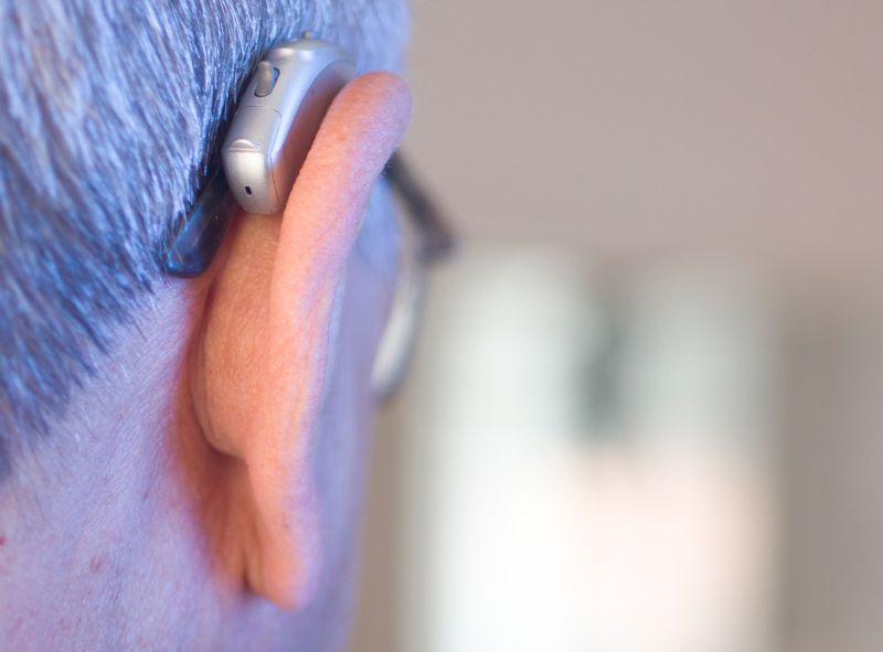 Close-up of a man's ear, showing a hearing aid and eyeglass arm resting over ear. 